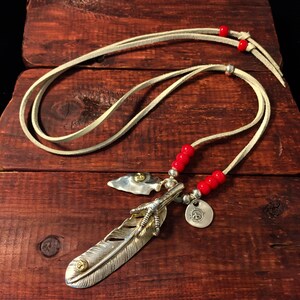 May include: A silver feather pendant necklace with a bird's foot charm and a small arrowhead charm. The necklace is on a white leather cord with red beads.