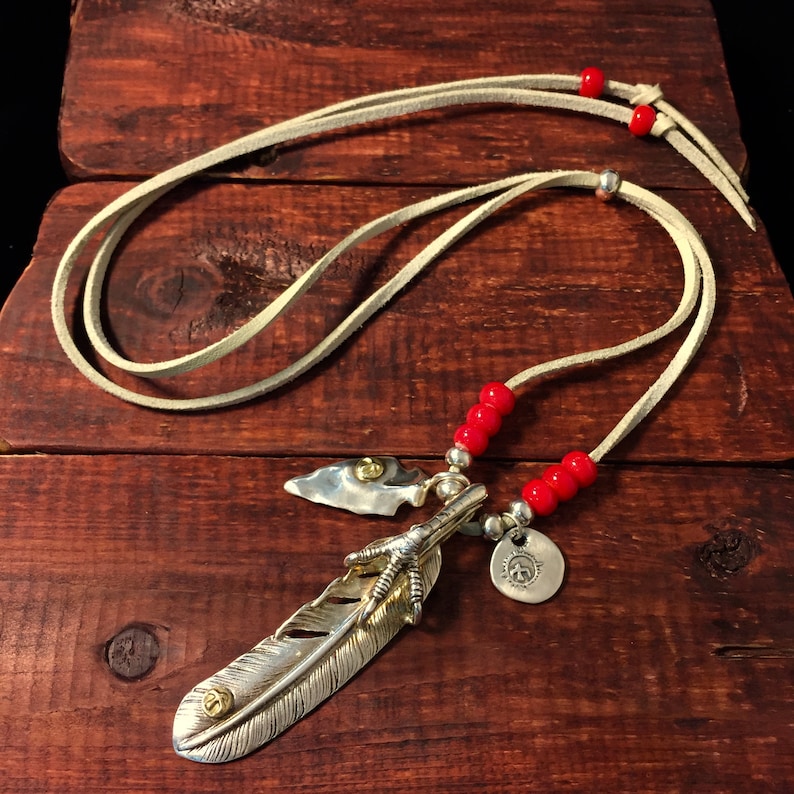 May include: A silver feather pendant necklace with a bird's foot charm and a small arrowhead charm. The necklace is on a white leather cord with red beads.
