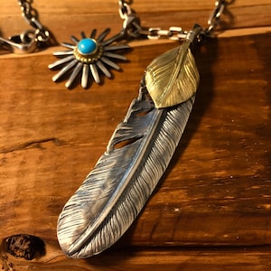 May include: A silver and gold feather pendant necklace with a turquoise stone accent. The feather is detailed with a textured design and the pendant is attached to a silver chain.