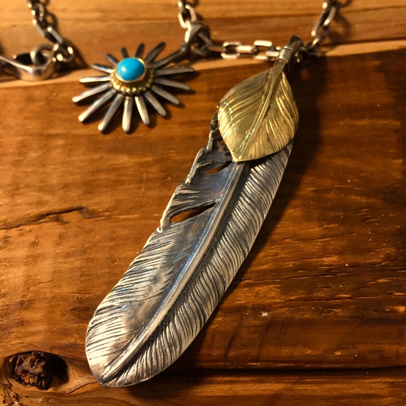 May include: A silver and gold feather pendant necklace with a turquoise stone accent. The feather is detailed with a textured design and the pendant is attached to a silver chain.