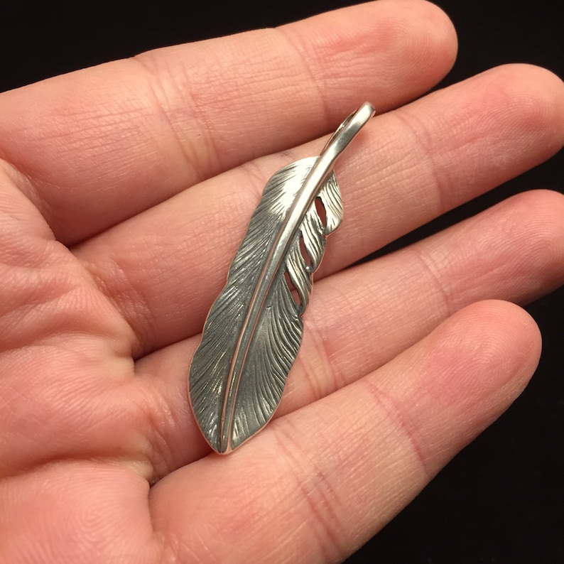 May include: A silver feather pendant with detailed veining and a loop at the top for a chain. The pendant is held in a hand against a dark background. The feather design is a popular motif for jewelry.