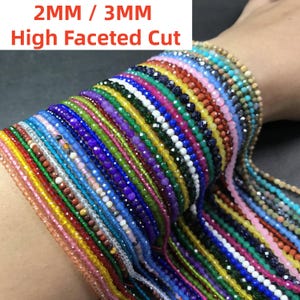 Puede incluir: Una colección de cuentas coloridas de corte facetado en varios colores, incluyendo rojo, azul, verde y morado. Las cuentas miden 2 mm y 3 mm, están ensartadas y se muestran en primer plano. El texto de la imagen dice "2MM / 3MM High Faceted Cut."