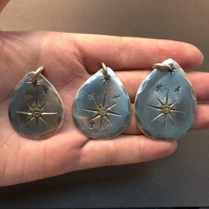 May include: Three silver teardrop-shaped pendants with a starburst design and a small gold dot in the center. Each pendant has a small loop at the top for attaching a chain.
