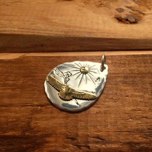 May include: A silver pendant with a gold eagle flying towards a sun with rays. The pendant is shaped like a teardrop and has a textured surface.