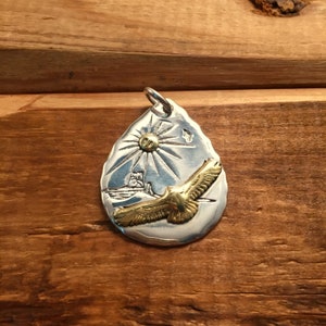 May include: Silver teardrop pendant with a gold eagle in flight, a sun with rays, and a small image of a lighthouse.