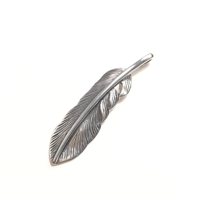 May include: A silver-colored feather pendant with detailed veining. The pendant is a stylized representation of a feather, with a loop at the top for a chain. The feather is set against a white background.