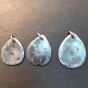 May include: Three silver pendant charms in the shape of a teardrop with a textured surface. Each charm has a small circle in the center and a few small dots around the circle.