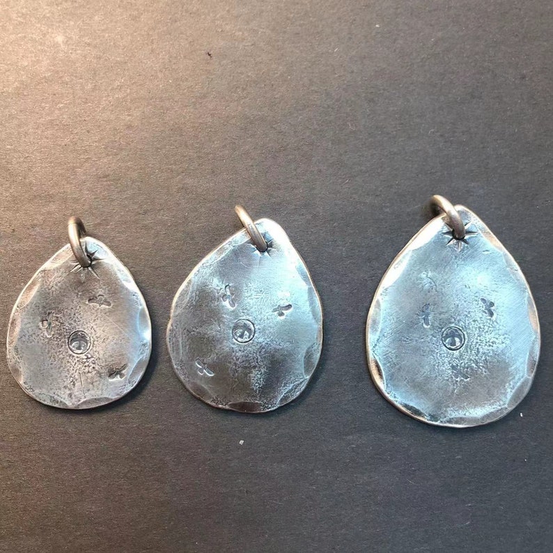 May include: Three silver pendant charms in the shape of a teardrop with a textured surface. Each charm has a small circle in the center and a few small dots around the circle.