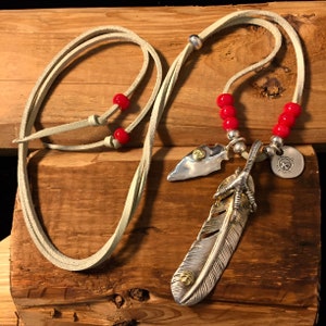May include: A silver feather pendant necklace with a bird's foot, a silver arrowhead, and red beads on a light brown leather cord.