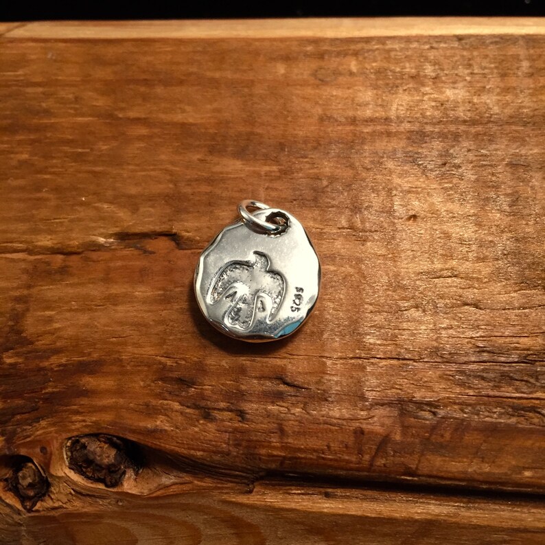 K&ouml;nnte beinhalten: Silberner Anh&auml;nger mit einem Vogelmotiv und der Gravur "SOS" auf der R&uuml;ckseite.