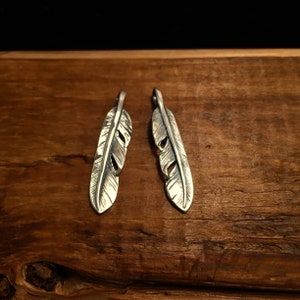 Puede incluir: Dos encantos de plumas de plata con grabado detallado, mostrados sobre una superficie de madera.