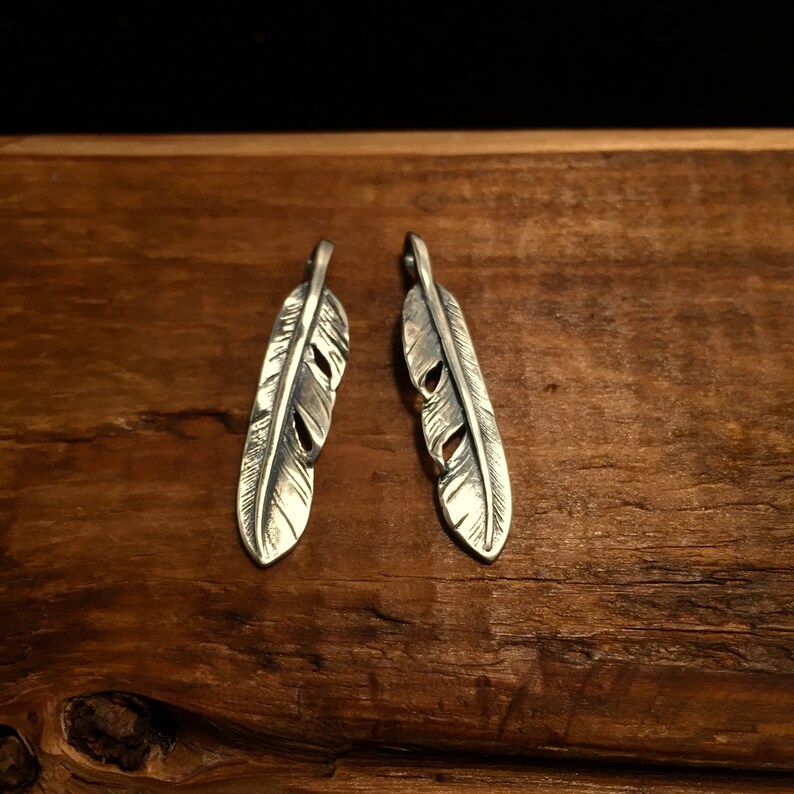 Puede incluir: Dos encantos de plumas de plata con grabado detallado, mostrados sobre una superficie de madera.