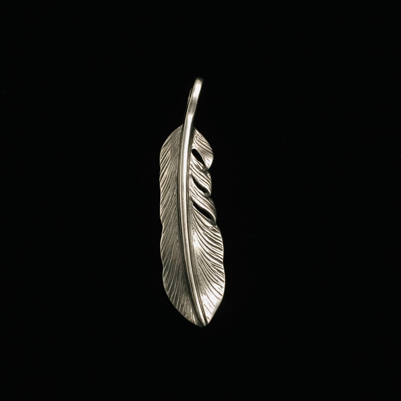 May include: A silver-colored feather pendant with intricate detailing. The pendant is suspended from a small loop at the top and features a realistic feather design with visible barbs and a central quill. The background is solid black.