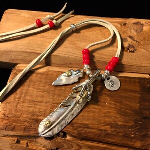 May include: A silver feather pendant necklace with a bird's foot charm, a small arrowhead charm, and red beads on a beige leather cord.