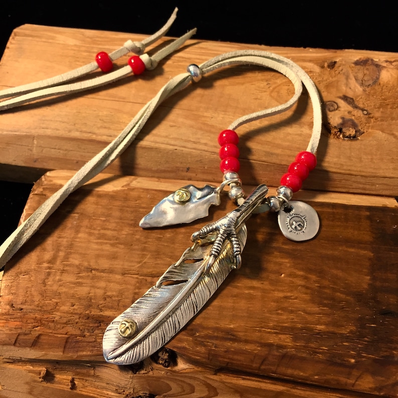 May include: A silver feather pendant necklace with a bird's foot charm, a small arrowhead charm, and red beads on a beige leather cord.