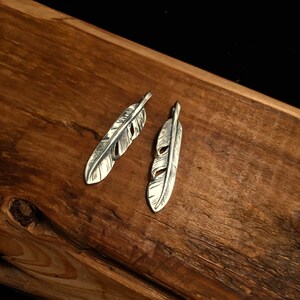Puede incluir: Dos encantos de plumas de plata con dise&ntilde;os recortados. Los encantos est&aacute;n sobre una superficie de madera.