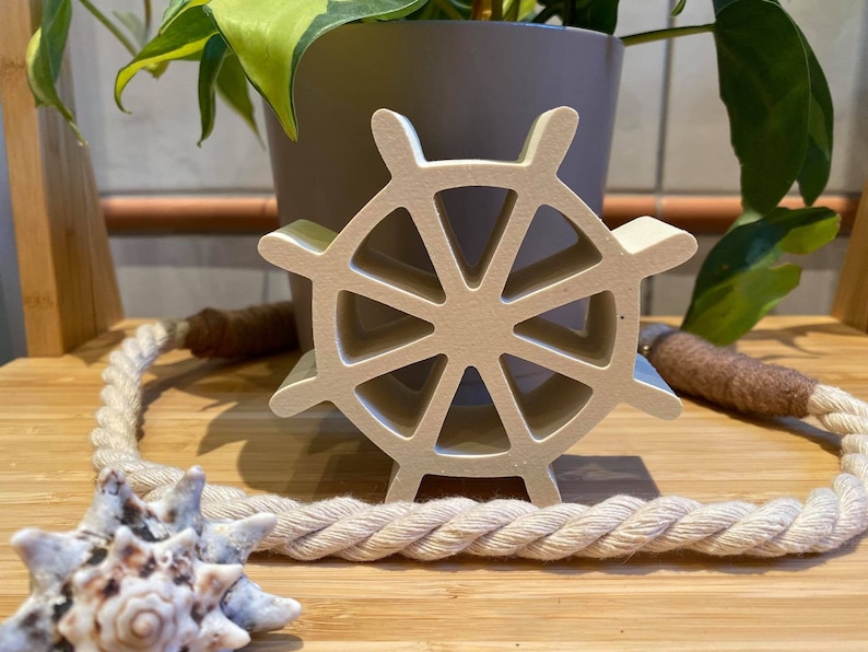 以下が含まれることがあります： 白い木製の船の舵輪にロープが巻かれている。舵輪は木製の表面に置かれ、手前に貝殻がある。