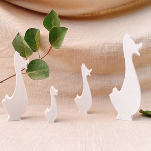 Könnte beinhalten: Vier weiße Gänsefiguren in verschiedenen Größen sind auf einer beigen Oberfläche angeordnet, mit einem Zweig grüner Blätter im Hintergrund. Die Gänse haben ein minimalistisches Design mit langen Hälsen und stilisierten Schnäbeln.