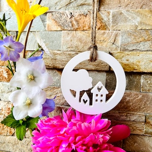 Könnte beinhalten: Weißer Holzausschnitt eines Hauses mit einem Baum und Herzen, die an einer Schnur an einer Steinmauer hängen. Das Haus hat Fenster und eine Tür. Der Baum hat eine wolkenartige Form. Der Ausschnitt ist von rosa, weißen und gelben Blumen umgeben.