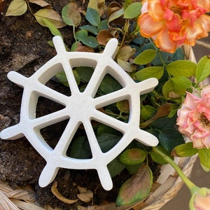 以下が含まれることがあります： 8本のスポークが付いた白いセラミック製の船の舵が、土と緑と茶色の葉で満たされた籐のバスケットの中に置かれています