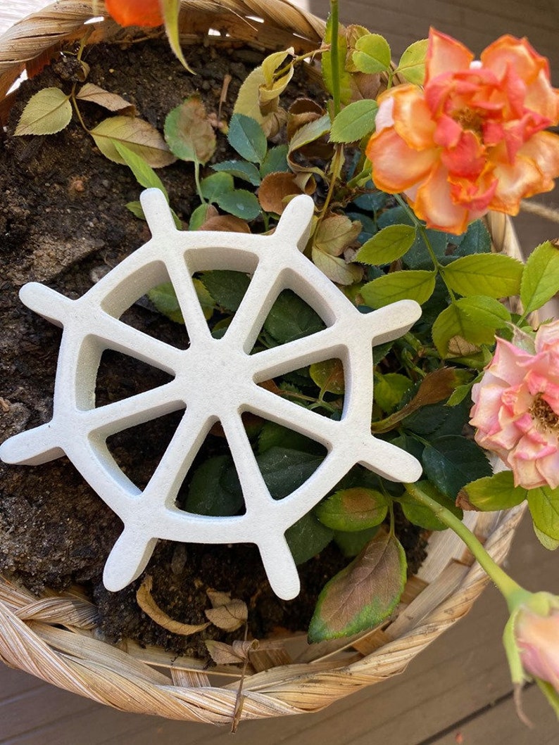 以下が含まれることがあります： 8本のスポークが付いた白いセラミック製の船の舵が、土と緑と茶色の葉で満たされた籐のバスケットの中に置かれています