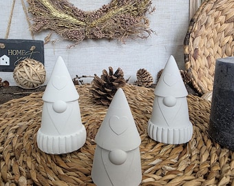 Puede incluir: Tres figuras de gnomos blancos con detalles de corazón, dispuestas sobre una estera tejida. Los gnomos tienen sombreros cónicos y narices redondas. Una corona decorativa y otros elementos naturales están en el fondo.