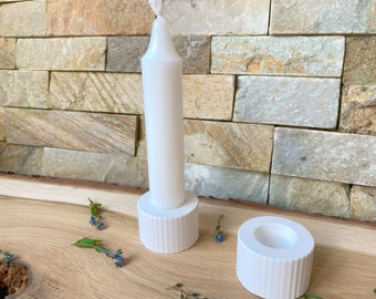 May include: A white candle stands in a white, ribbed candle holder. Another matching holder sits nearby. The candle and holders are on a wooden surface, with a stone wall in the background. Small blue flowers are scattered around.