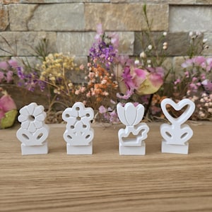 Könnte beinhalten: Vier kleine, weiße Dekorationsgegenstände in Form von Blumen und einem Herz. Jeder Gegenstand steht auf einem kleinen, rechteckigen Sockel. Der Hintergrund zeigt eine Vielzahl von Trockenblumen in Rosa, Lila, Orange und Gelb.