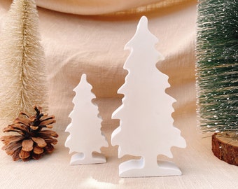 May include: Two white, stylized Christmas tree figurines of different sizes. A small pine cone and two miniature Christmas trees are also in the image. The background is a neutral beige fabric, creating a festive holiday scene.