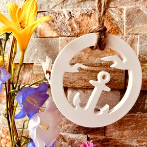 Könnte beinhalten: Ein weißer Holz-Kreis mit einem Ausschnitt eines Ankers und Möwen. Der Kreis hängt an einem braunen Seil.