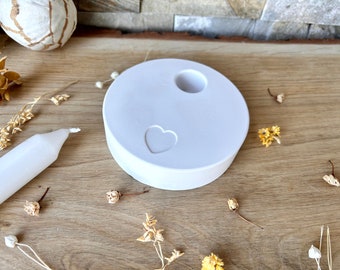 May include: White ceramic candle holder with a heart cutout and a small hole in the center for a candle. The holder is on a wooden surface.