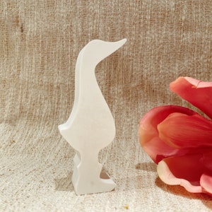 Könnte beinhalten: Weißer Holzschnitt einer Gans oder Ente, die auf einem beigefarbenen Stoffhintergrund steht, mit einer rosa Magnolienblüte im Hintergrund.