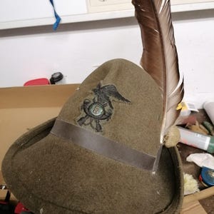 Könnte beinhalten: Ein olivgrüner Filzhut mit breiter Krempe und einer langen, braunen Feder an der Seite. Auf der Vorderseite befindet sich ein dunkles, gesticktes Emblem eines Vogels, der über einem runden Abzeichen mit der Zahl 5 thront.