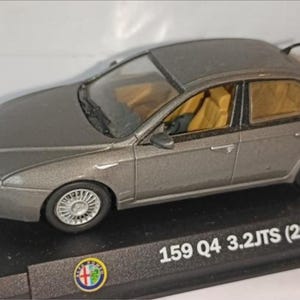 Peut inclure: Modèle réduit gris d'une Alfa Romeo 159 Q4 3.2 JTS (2005). La voiture miniature est posée sur un socle noir avec le nom et l'année de la voiture en blanc. L'intérieur est beige et les roues sont argentées.