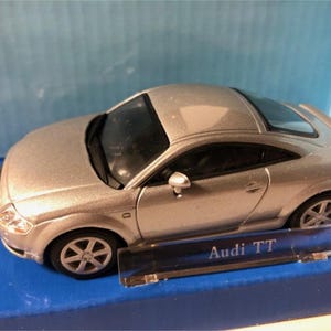 Può includere: Modellino di auto Audi TT argentata con ruote nere e parabrezza nero. Il modello è appoggiato su una superficie blu e presenta la scritta "Audi TT" su una base di plastica trasparente.