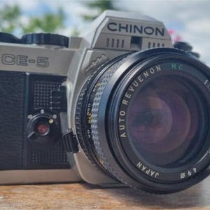 May include: A silver and black Chinon CE-5 film camera with an Auto Revuenon lens. The lens has a 50mm focal length and f/1.4 aperture. The camera has a black strap attached and is sitting on a wooden surface.