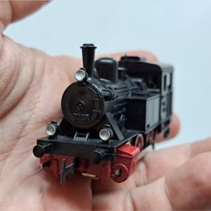 Puede incluir: Un modelo de tren en miniatura negro y rojo, con un diseño detallado que incluye una chimenea, faros y una cabina. El tren de juguete se sostiene en una mano, destacando su pequeño tamaño y sus intrincadas características. Un juguete clásico para los entusiastas de los trenes.