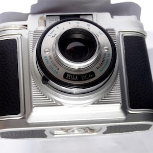 Könnte beinhalten: Eine Vintage Bella DC4 Kamera in Silber und Schwarz. Die Kamera hat einen schwarzen Ledergriff auf jeder Seite und ein Objektiv mit Fokusring. Das Objektiv trägt die Aufschrift "B&W COLOR" und "BELLA DC4".