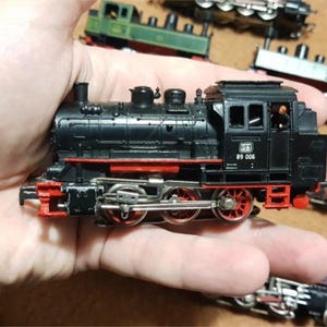 May include: A black and red model train with the number 89 006 on the side. The model train is held in a hand, with other model trains in the background. The train has red wheels and accents.