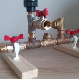 May include: A plumbing system with multiple valves and pipes, mounted on two wooden boards. The valves have red handles and are connected to a silver pipe. The system is on a light brown wooden surface.