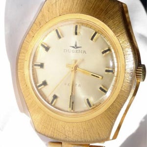 Könnte beinhalten: Eine goldfarbene Vintage-Armbanduhr mit strukturierter Lünette und Armband. Das Zifferblatt ist cremefarben mit schwarzen Markierungen und Zeigern. Der Markenname "DUGENA" ist oben mittig mit "FETA" darunter abgebildet. Klassisches Retro-Design.