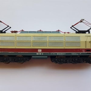 Puede incluir: Una maqueta de locomotora eléctrica en crema, rojo y verde. El modelo de tren tiene dos pantógrafos, múltiples ventanas y componentes detallados del tren de rodaje. La locomotora es una réplica de una DB 103 118-3.