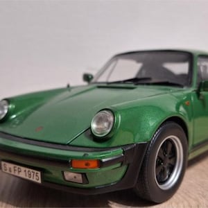 May include: A green model car with black wheels and a spoiler. The car has a license plate that reads "S FP 1975". The car is on a wooden surface.