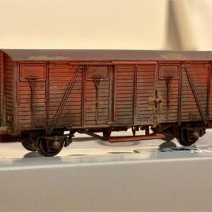 Op de afbeelding: Gedetailleerd model van een vintage goederenwagon, met een verweerde, roestbruine buitenkant. Het model heeft zichtbare deuren, wielen en ingewikkelde details, wat duidt op een historisch of verzamelobject. De wagon staat op een lichtgekleurd oppervlak.