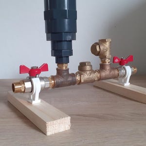 May include: A plumbing assembly featuring a dark gray cylindrical component connected to brass and copper pipes. Red-handled valves and white supports are visible. The assembly is mounted on wooden blocks.