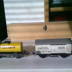 Puede incluir: Dos vagones de tren en miniatura. Uno es un vagón cisterna amarillo Shell con el logotipo de Shell. El otro es un vagón cubierto blanco Amstel Bier. Ambos están sobre una vía. Una caja de madera está en el fondo.