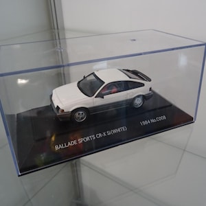 Puede incluir: Un coche modelo blanco y negro, un Ballade Sports CR-X Si (White), se exhibe en una caja de plástico transparente. El coche modelo está sobre una base negra con el texto blanco "1984 No.C008".