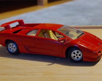 Può includere: Un'auto giocattolo rossa con spoiler e ruote argentate. L'auto ha un design elegante con un profilo ribassato e interni gialli. L'auto giocattolo è una replica in miniatura di un'auto sportiva.