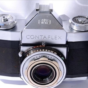 Puede incluir: Una cámara Contaflex vintage con cuerpo plateado y detalles en cuero negro. La lente tiene múltiples anillos con números y texto. La cámara tiene el texto "CONTAFLEX" y "ZEISS IKON".