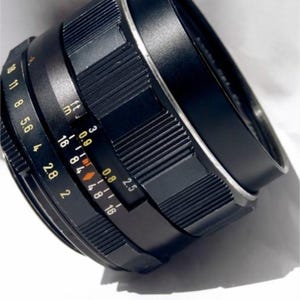 May include: A black camera lens with a silver ring and markings in yellow and orange. The lens has a 2.5 aperture and a range of 16 to 84 millimeters.